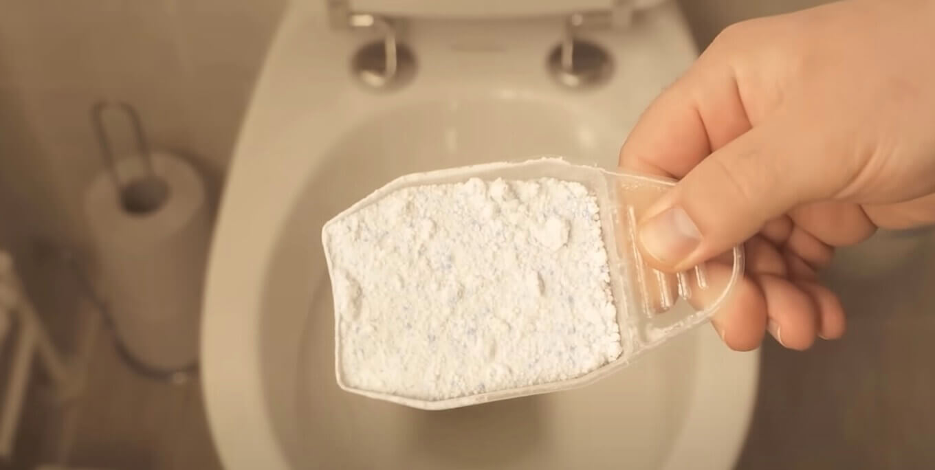 Hand holding a scoop of white laundry detergent powder over a toilet bowl