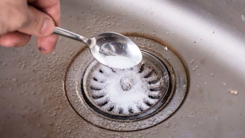 Hand pouring white powder into a kitchen sink drain with a spoon